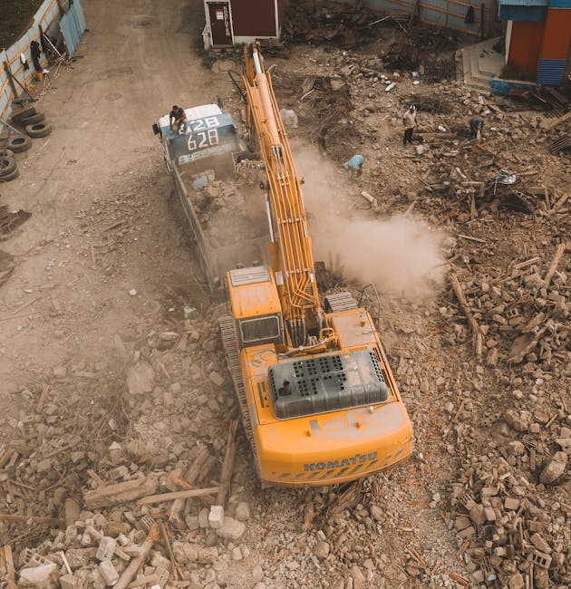 An overhead view of a construction site showing a large yellow excavator with a long arm and bucket attachment clearing debris from an area filled with broken bricks, concrete, and rubble. The excavat